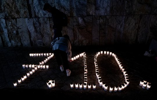 Brennende Grablichter bilden auf nächtlichem Kopfsteinpflaster das Datum „7.10.“, eine Person in gestreiftem Pullover stellt eine weitere Kerze dazu.