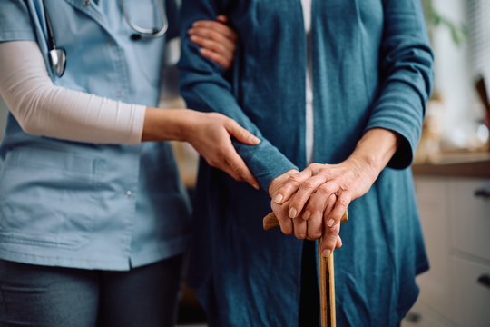 Eine Pflegekraft mit Stethoskop stützt den Arm einer älteren Person, die sich mit beiden Händen auf einen Gehstock aus Holz stützt.