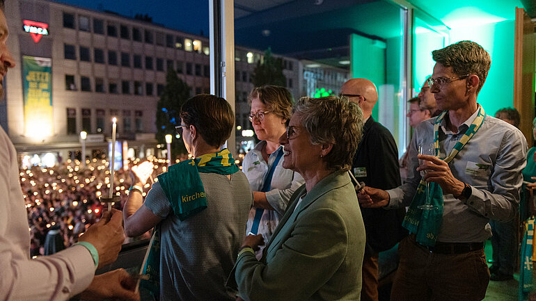 Fraktionsvorsitzende Britta Haßelmann steht vor einem Fenster, hinter dem zahlreiche Menschen mit Kerzen zu sehen sind.