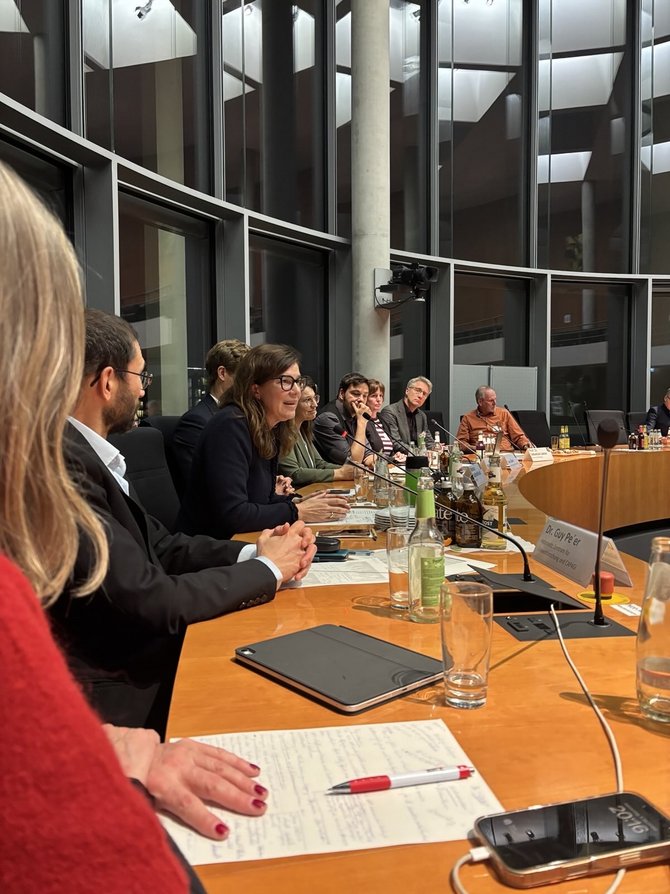 Innenaufnahme eines modernen Sitzungssaals mit gebogener Tischreihe; mehrere Personen sitzen dicht nebeneinander, sprechen in Mikrofone, vor ihnen stehen Wasser- und Getränkeflaschen sowie Notizblöcke, im Vordergrund ist eine Person mit rotem Pullover und Notizblock zu sehen, im Hintergrund spiegeln sich Decke und Licht in hohen Glasfenstern.