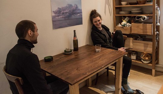 Eine junge Frau und ein junger Mann sitzen sich in einer spärlich eingerichteten Küche gegenüber