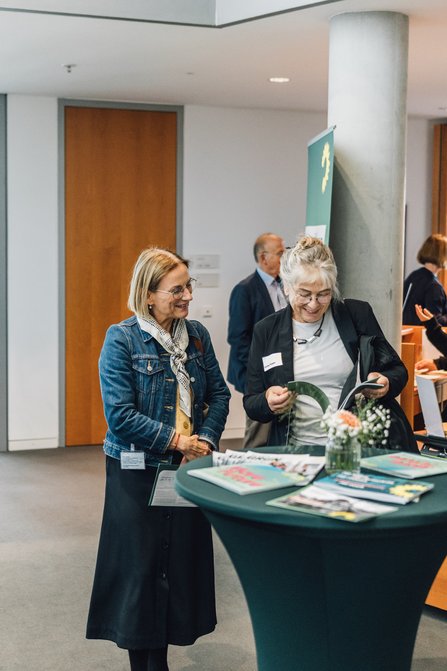 Das Bild zeigt 2 Personen an einem Stehtisch, auf dem sich Publikationen und florale Dekoration befinden