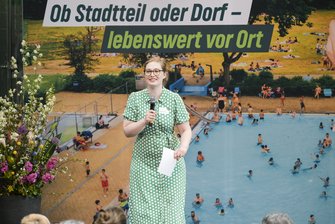 Eine Frau in Kleid mit floralem Muster steht auf der Bühne, im Hintergrund das Veranstaltungsmotiv - ein Freibad.