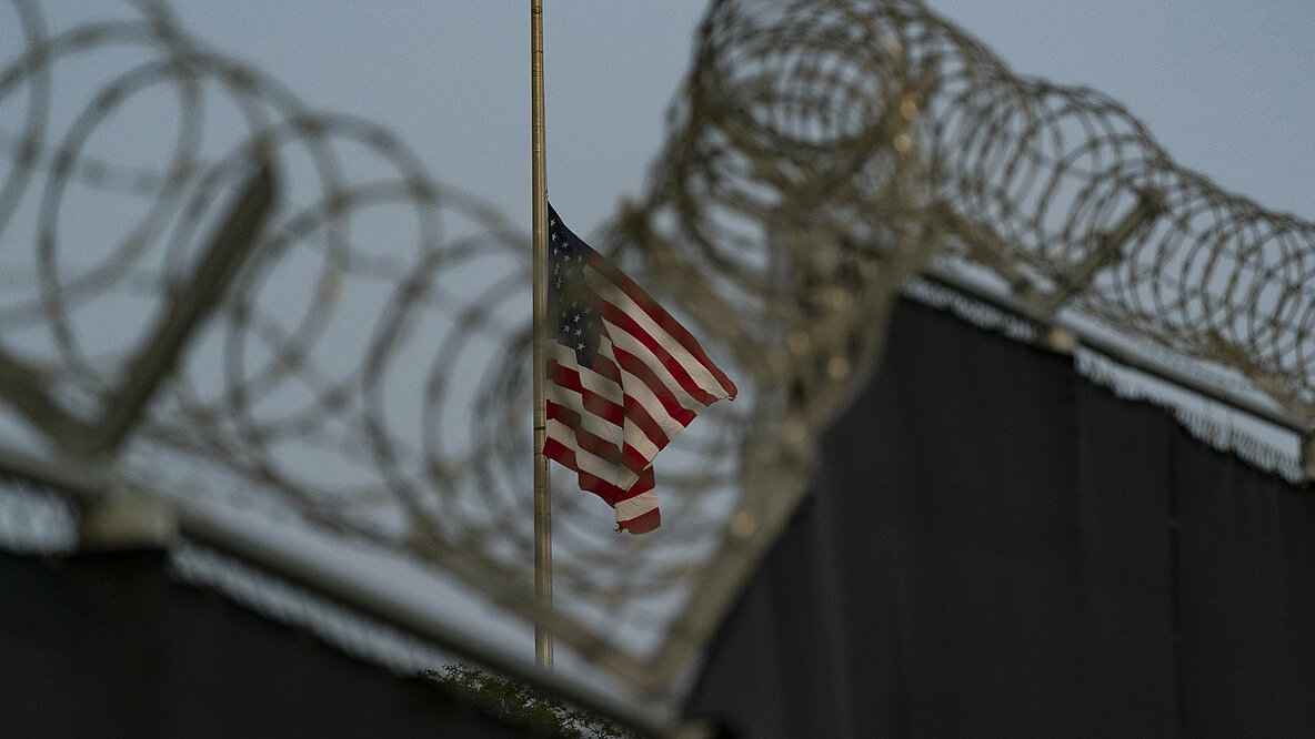 Eine Mauer, oben drauf Stacheldraht im Hintergrund die US-Flagge