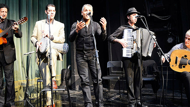 Fünf Männer mit Musikinstrumenten stehen auf einer Bühne mit Mikrofonen. 