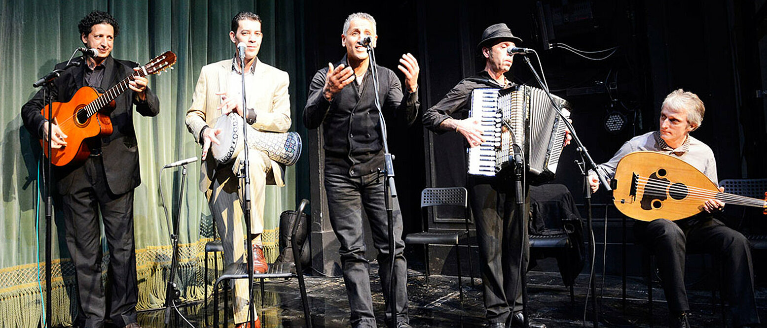 Fünf Männer mit Musikinstrumenten stehen auf einer Bühne mit Mikrofonen. 