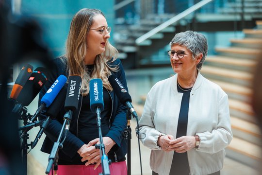 Katharina Dröge und Britta Haßelmann stehen hinten Mikrofonen und lächeln sich an