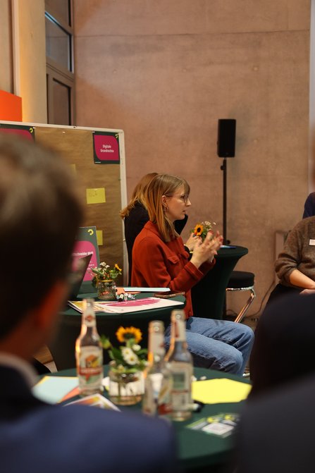Frau mit Mikro im Profil, links eine Pinwand mit Moderationskarten, im Vordergrund zwei Personen mit Rücken