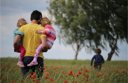 Ein Vater mit zwei Kindern auf dem Arm läuft über eine Wiese