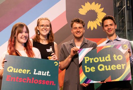 Vier Gäste auf der Bühne, zwei Schilder hochhaltend: ein Schild mit Queer.laut.entschlossen. Das andere mit Proud to be queer.